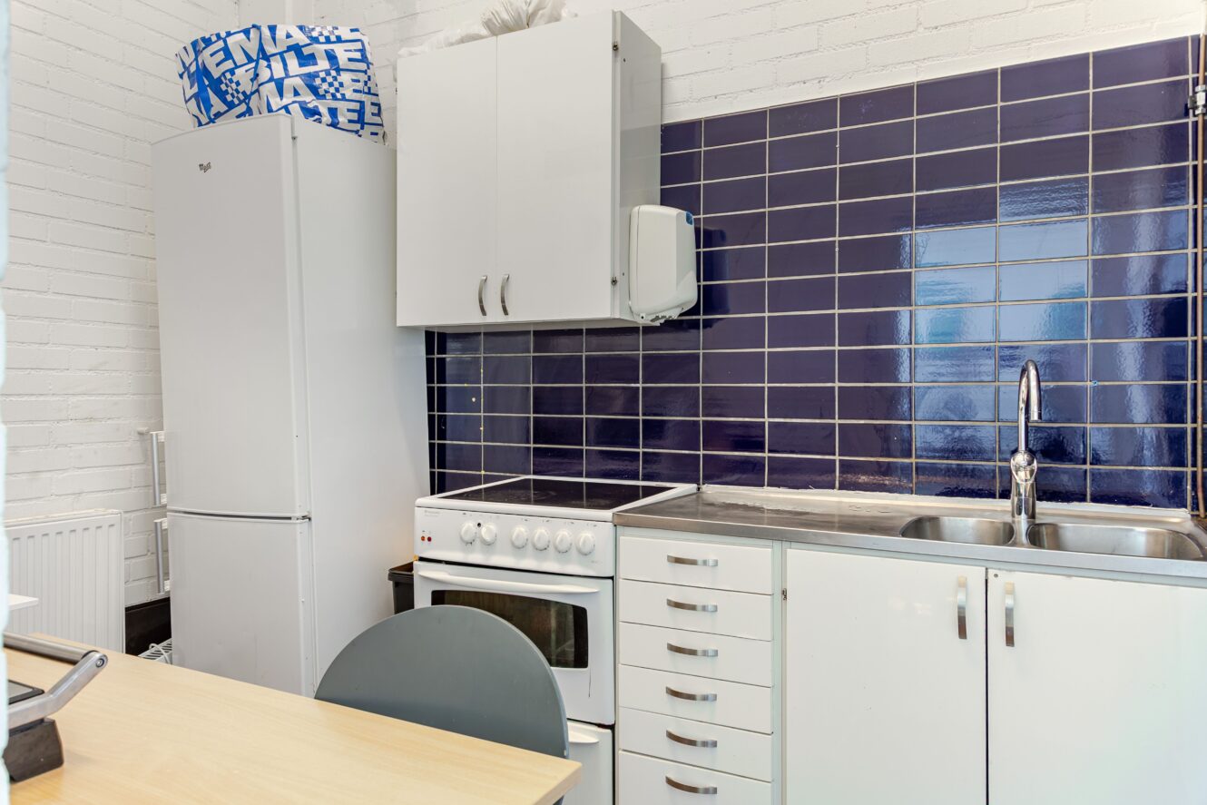 Litet kök med vita skåp, kylskåp, elspis, blå kaklade stänkskydd och en diskho i rostfritt stål. Blå och vita lådor är staplade ovanpå kylskåpet.