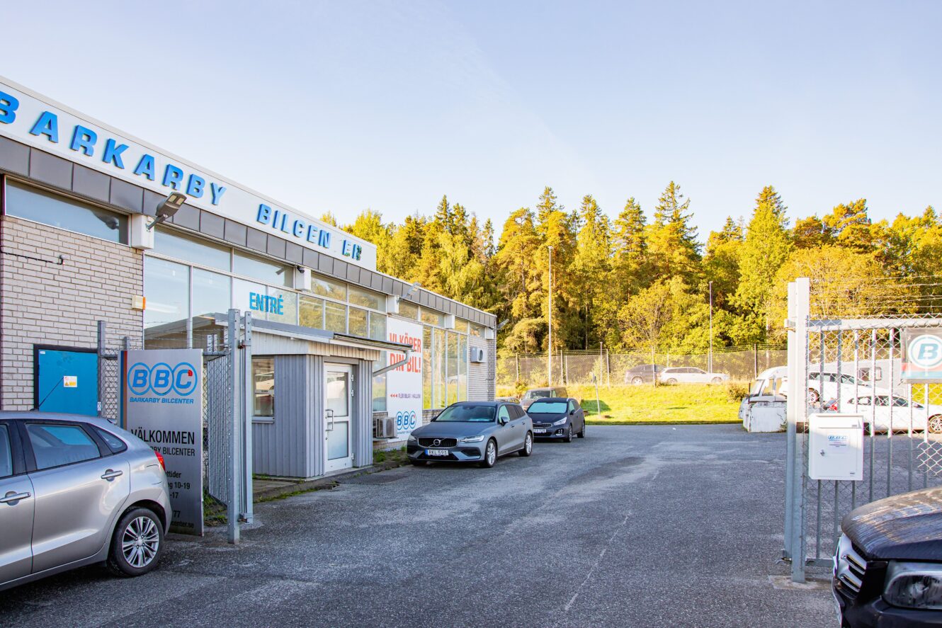 En parkeringsplats utanför en bilhandlare och verkstad med flera parkerade bilar, en inhägnad grind och träd i bakgrunden under en klar himmel.