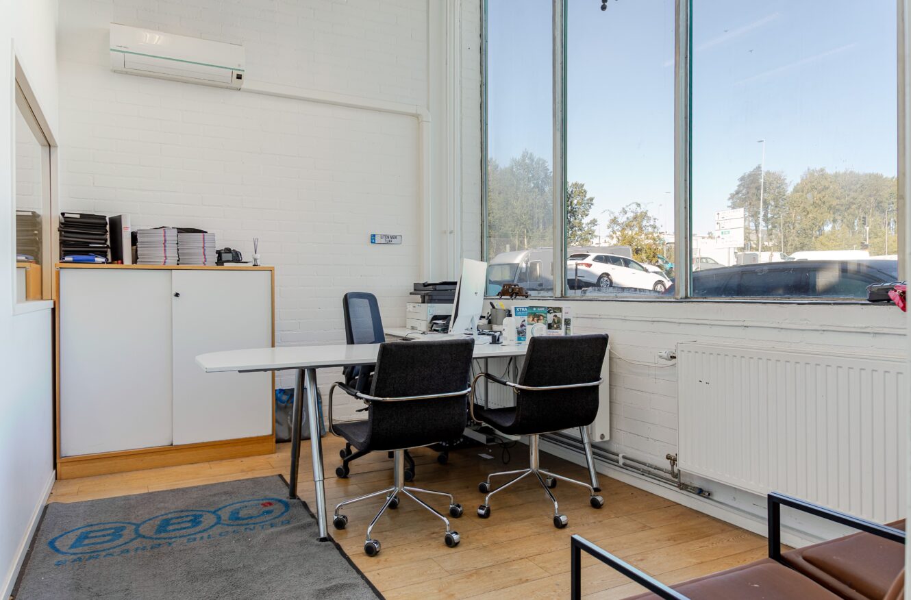 Ett litet kontor med ett vitt skrivbord, två svarta stolar, en dator, arkivskåp, stora fönster och en logomatta på trägolvet.