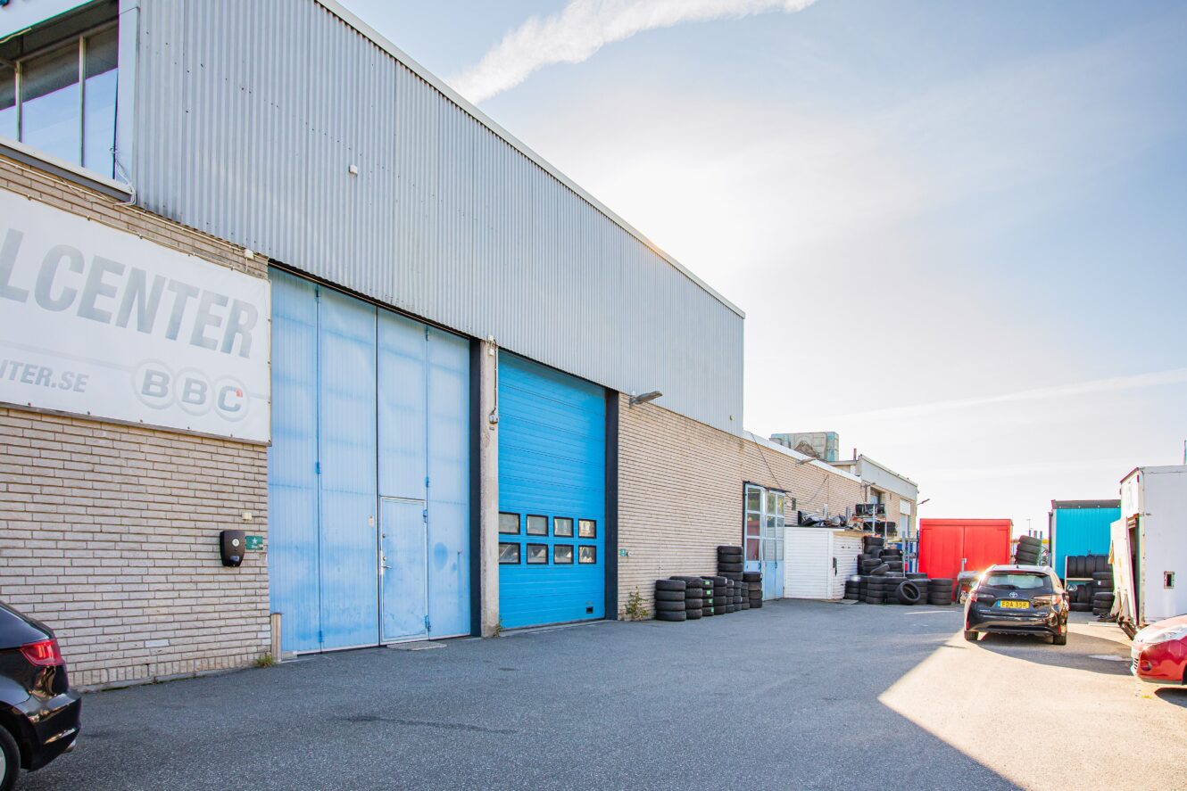 Industribyggnad med stora blå dörrar, däckstaplar, parkerade bilar och klar himmel i bakgrunden.