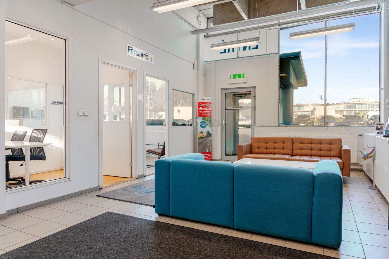 Ett modernt väntrum på ett kontor med en tealfärgad soffa, en brun soffa, klinkergolv och angränsande rum med glasväggar och kontorsstolar. Naturligt ljus kommer in genom stora fönster.