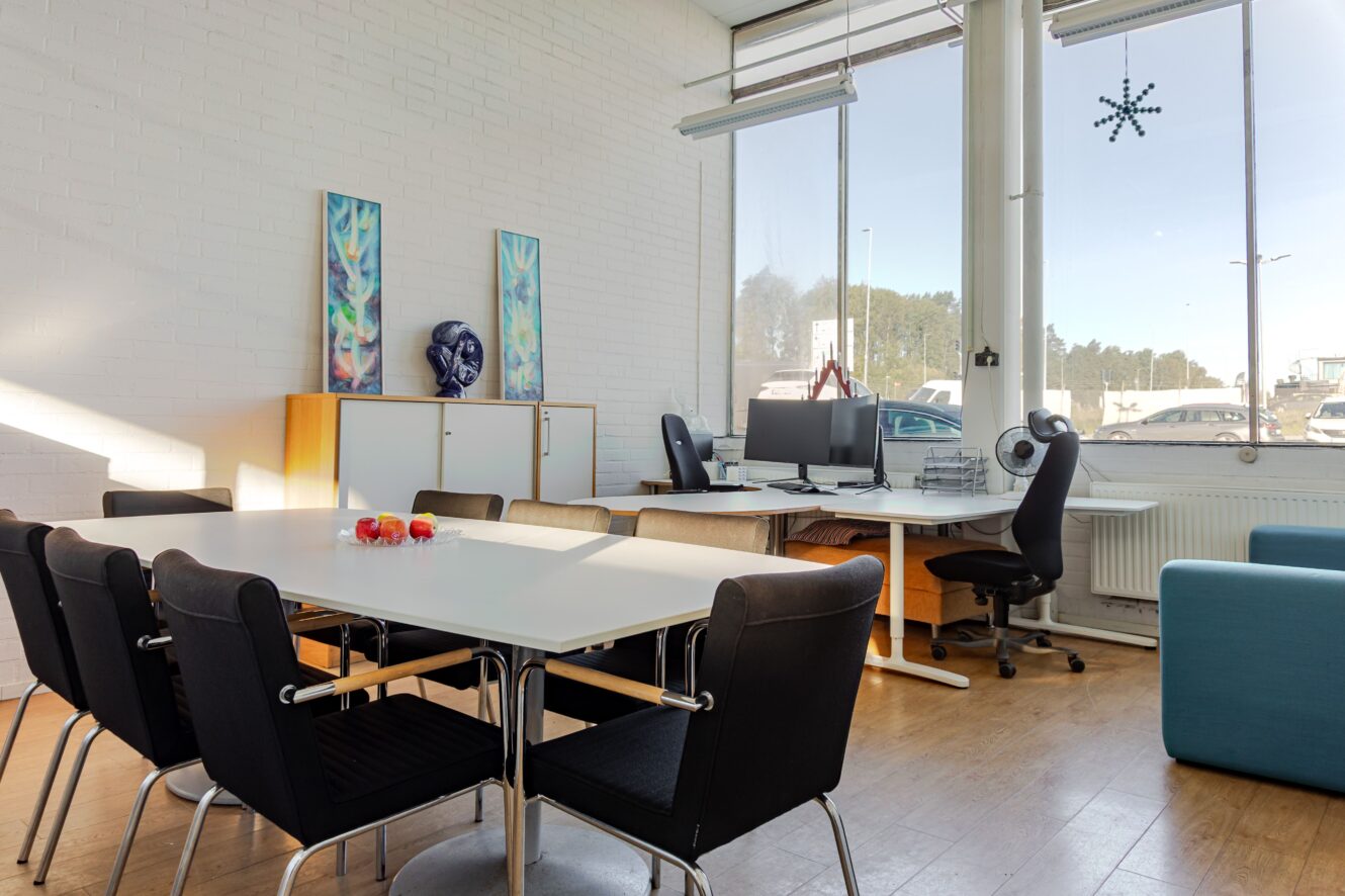 Modernt kontorsrum med mötesbord, svarta stolar, två skrivbord med bildskärmar vid stora fönster, konstverk på väggen och naturligt ljusinsläpp.