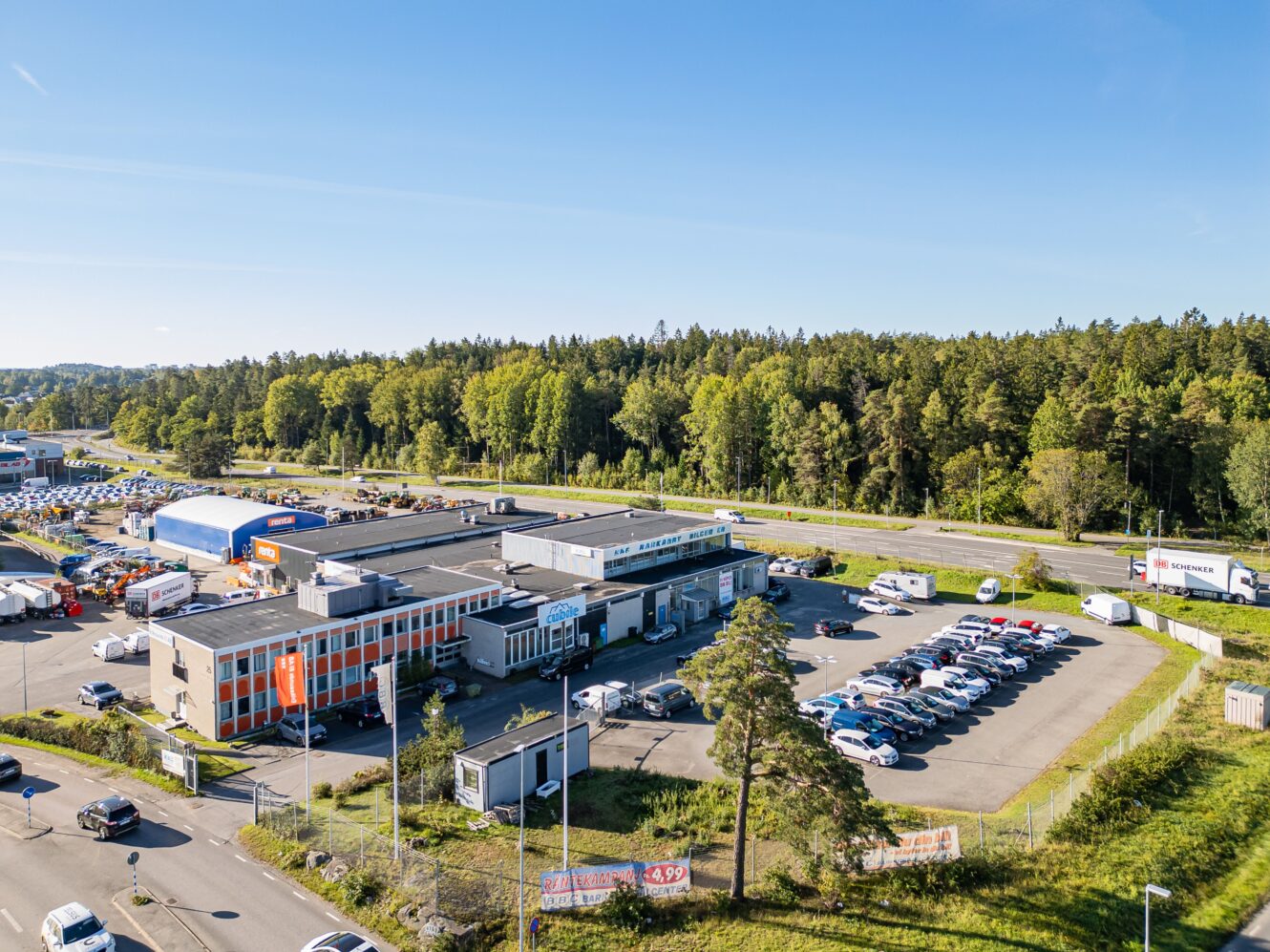 En kommersiell byggnad och en parkeringsplats syns nära en motorväg, omgiven av träd och grönska under en klarblå himmel.