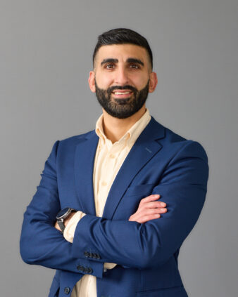 En man med skägg i blå kostym och beige skjorta står med armarna i kors och ler mot en grå bakgrund.