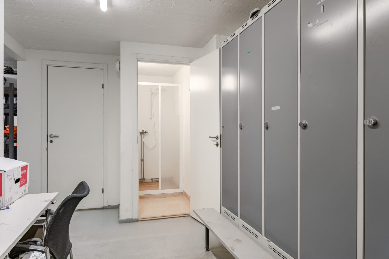 Ett omklädningsrum med gråa skåp, en bänk, ett skrivbord med en stol och en öppen dörr som leder till ett duschutrymme.