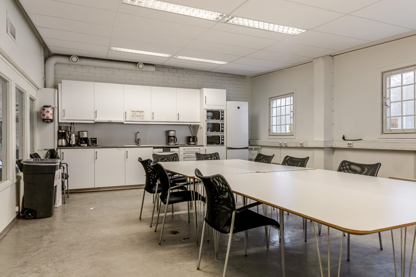 Personalens fikarum med vita bord, svarta stolar, pentry med skåp, kaffebryggare, mikrovågsugn, papperskorg och lysrör i taket.