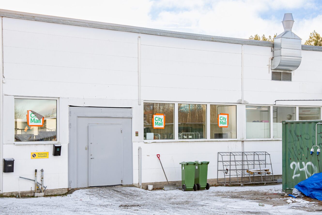 Bakre ingången till en City Mart-byggnad med gröna papperskorgar, ett metallställ och en grön container på en snöig mark.
