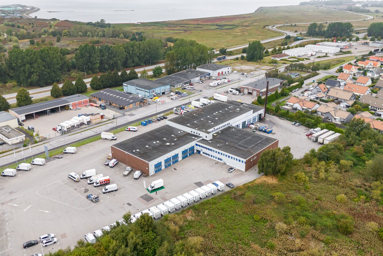 Flygfoto över en industrianläggning med flera lagerlokaler, parkerade lastbilar och närliggande bostadshus, nära ett vattendrag och ett grönt landskap.