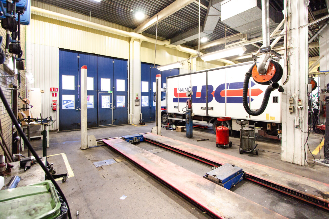 En lastbil står parkerad i ett väl upplyst garage för fordonsunderhåll med utrustning, verktyg och en hydraulisk lyft synlig.