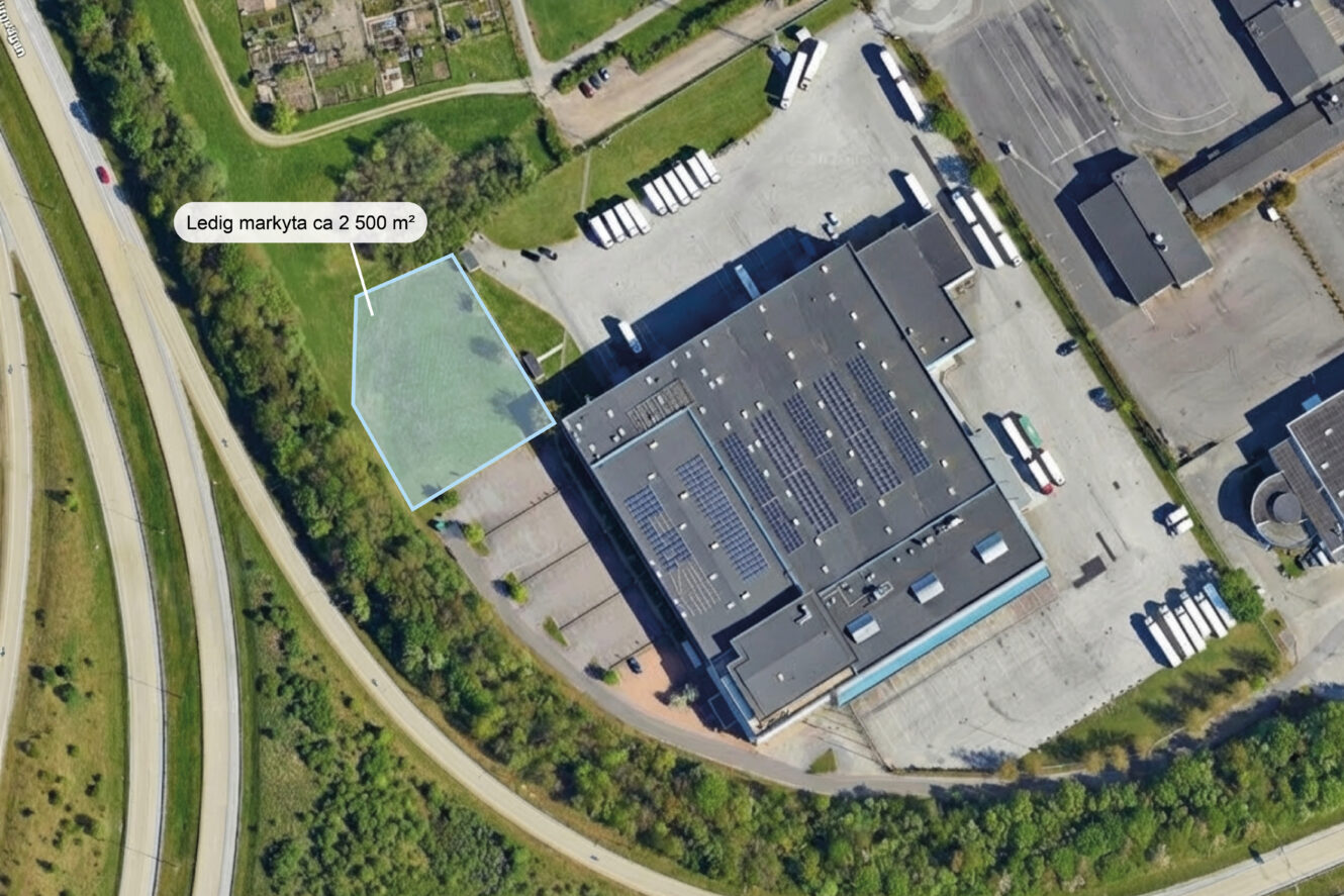 Flygfoto över en industribyggnad med en markerad ledig tomt i närheten, intill en väg och flera parkerade lastbilar.
