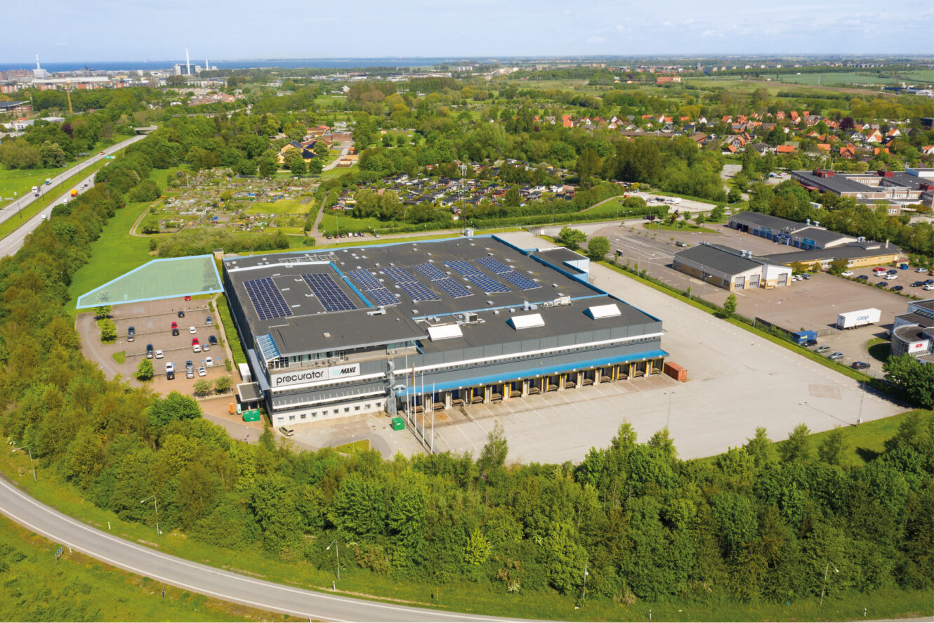 Flygfoto över en stor industrihall med solpaneler på taket, omgiven av parkeringsplatser, vägar och grönska.