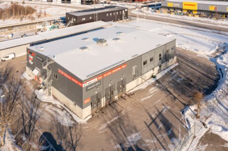 En stor grå industribyggnad med flera skyltar och dörrar, omgiven av snö och en asfalterad tomt, belägen nära andra kommersiella byggnader och en väg.