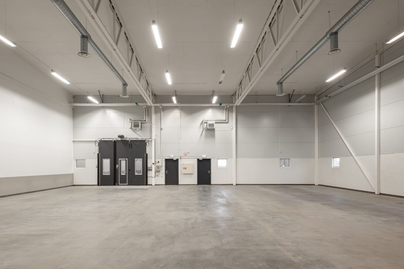 Rymlig, tom industrilagerlokal med högt i tak, betonggolv, vita väggar och flera synliga dörrar och ventilationskanaler.