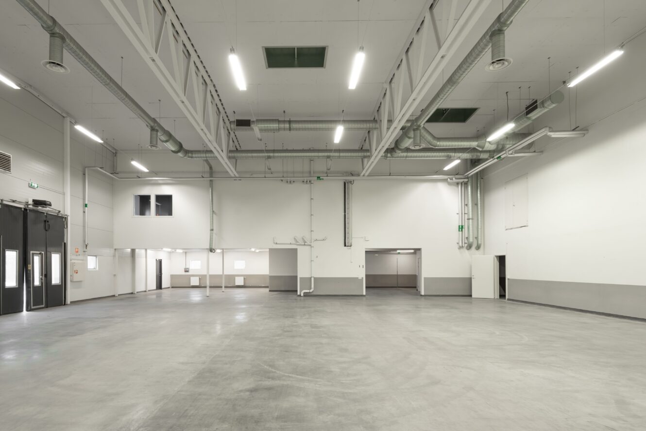 Stort tomt industrilager med vita väggar, grått golv, luftkanaler och starka taklampor. Flera dörrar och små fönster är synliga längs väggarna.