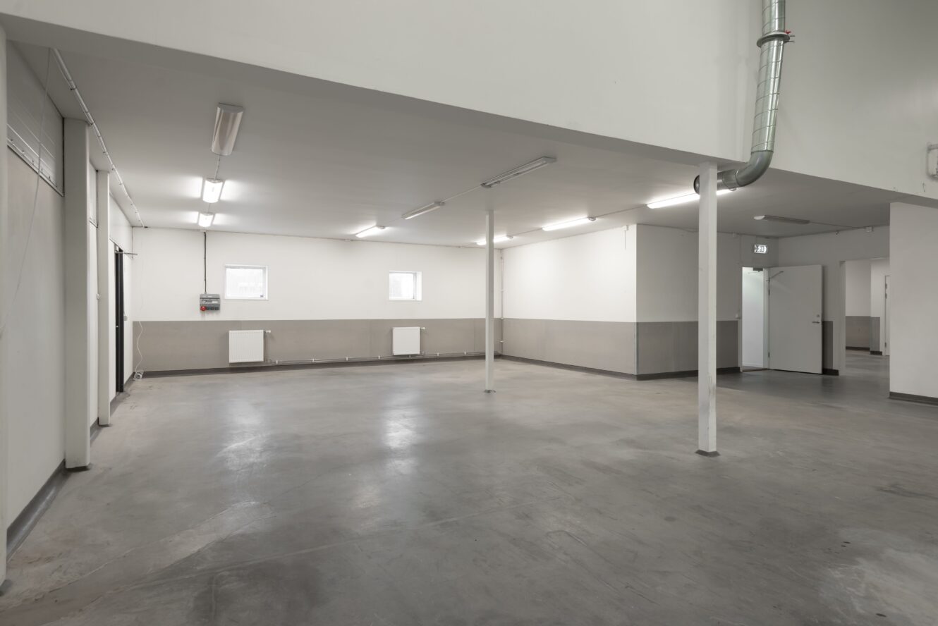Ett tomt, rent industrirum med betonggolv, vita väggar, flera pelare, taklampor, radiatorer, ventilationskanal och en dörr till höger.