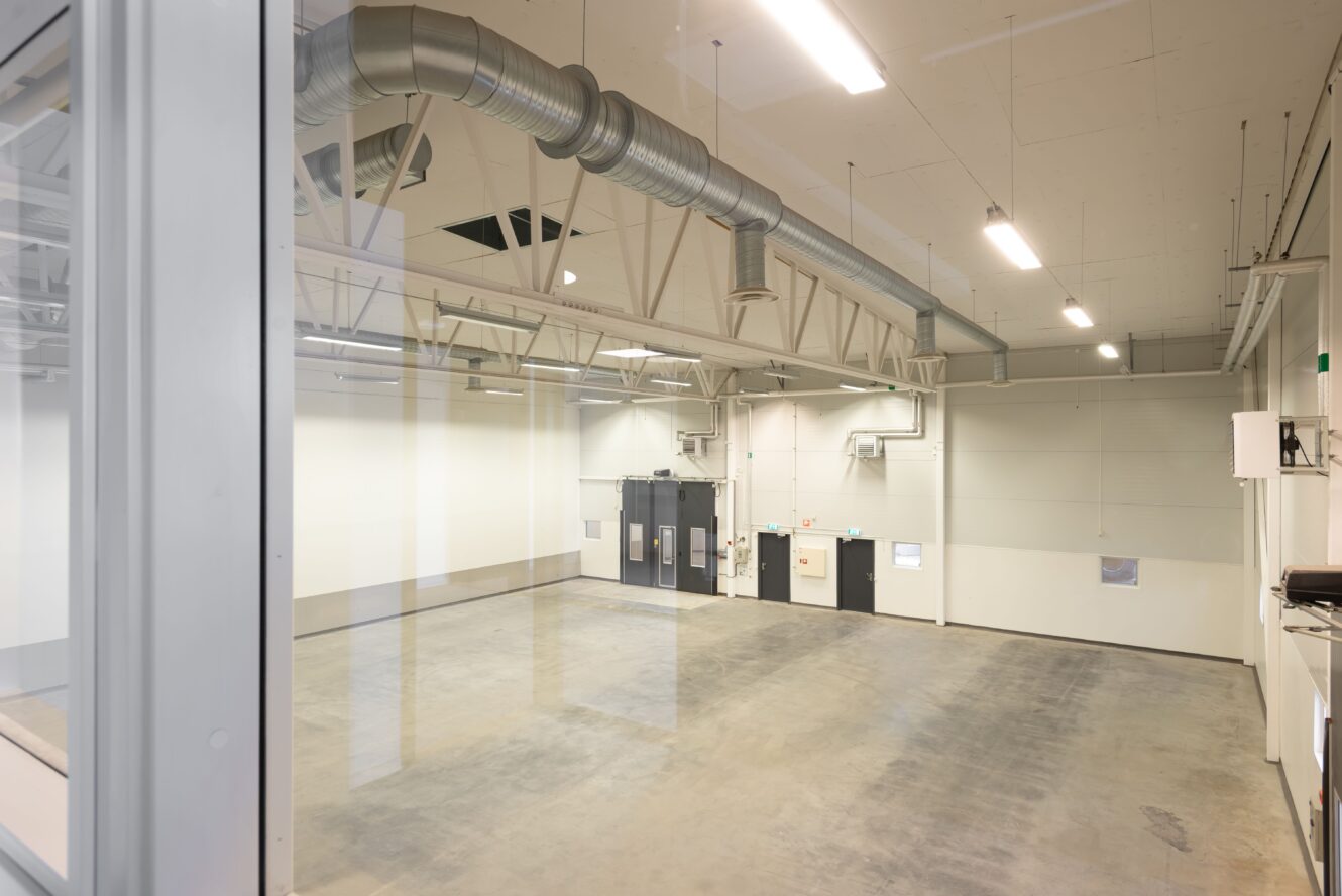 Tomt industrilager med polerat betonggolv, synliga takkanaler och lysrörsbelysning, sett genom ett glasfönster. Dubbeldörrar och elpaneler syns på den bortre väggen.