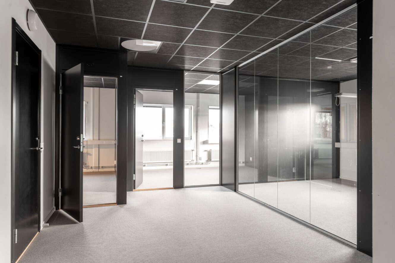 Tomt modernt kontorsutrymme med glasväggar, svarta dörrar och grå matta, med stora fönster som släpper in naturligt ljus.