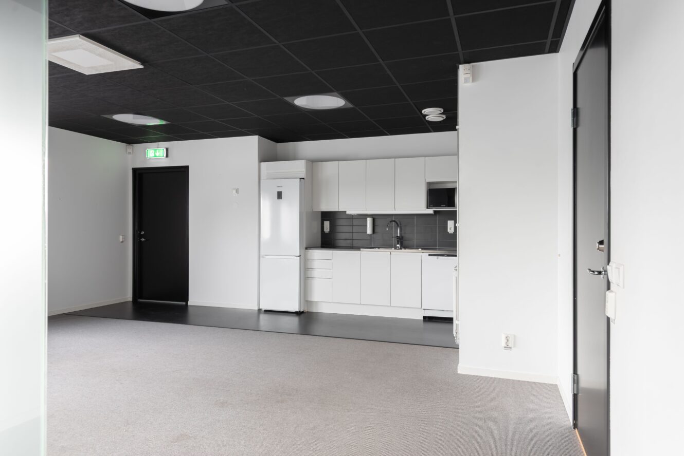 Ett modernt, omöblerat rum med ett litet vitt pentry, svartvita dörrar, grå heltäckningsmatta och svart klinkergolv under köksdelen.