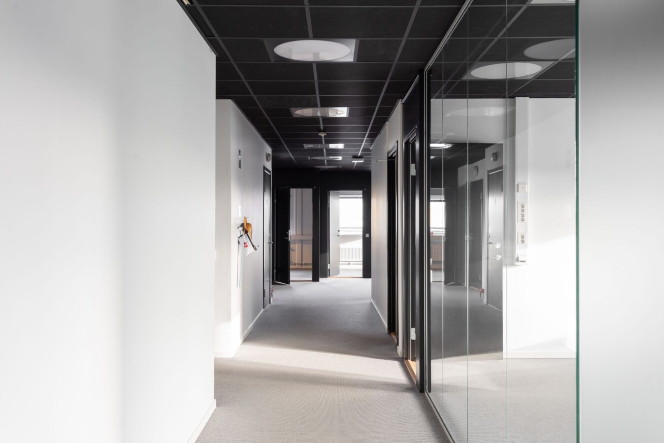 En modern, tom kontorskorridor med vita väggar, svart tak, grå matta och glaspaneler som reflekterar korridoren. Dörrar till kontor syns längs båda sidor.