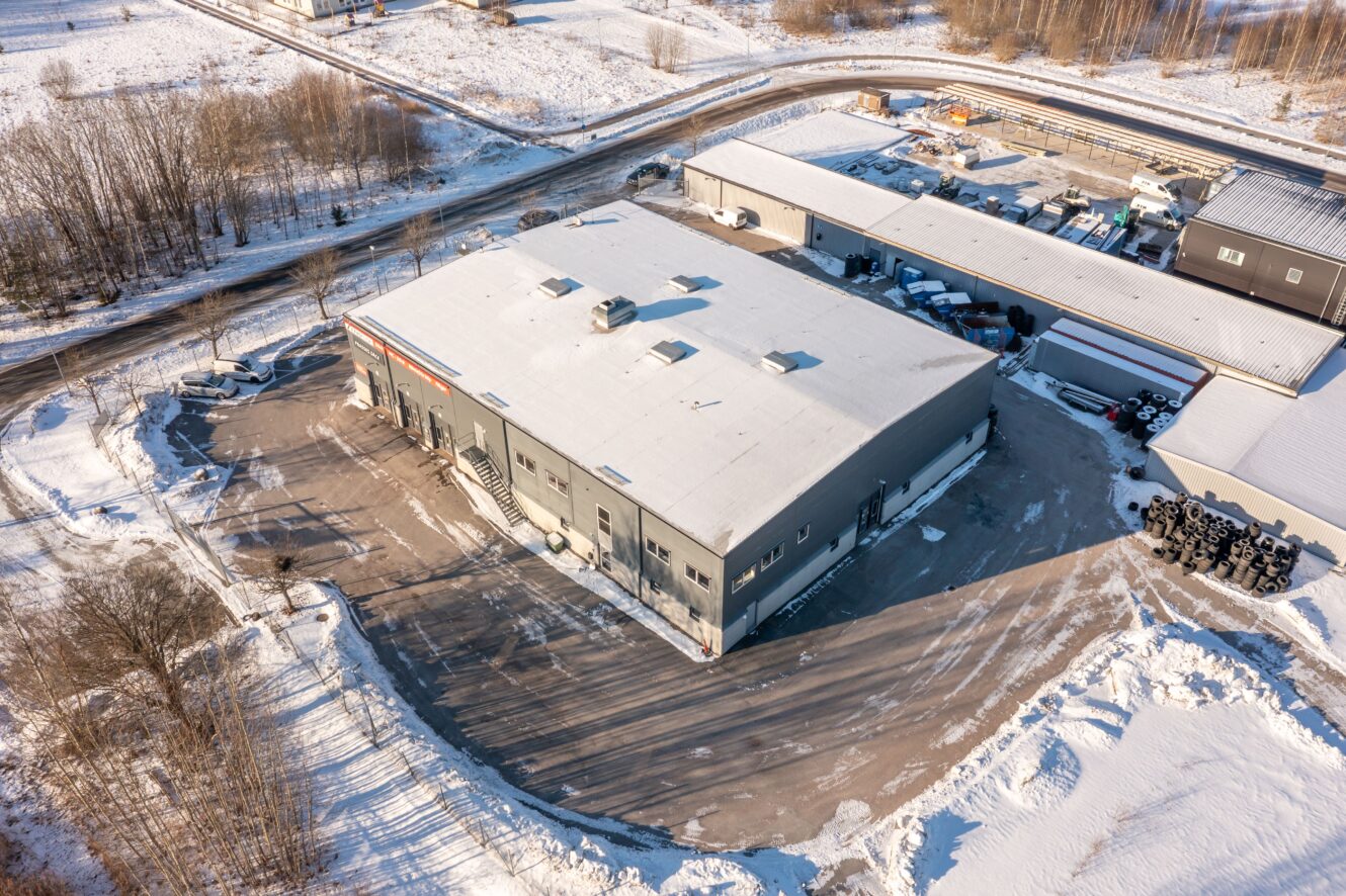 Flygfoto över ett stort industriellt lager med snötäckt tak och omgivningar, beläget intill andra byggnader och en röjt parkeringsplats.