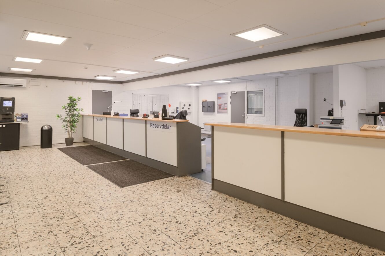 Receptionsområde med en lång disk, datorstationer, kontorsstolar, krukväxt och klinkergolv; vita väggar och tak med taklampor.