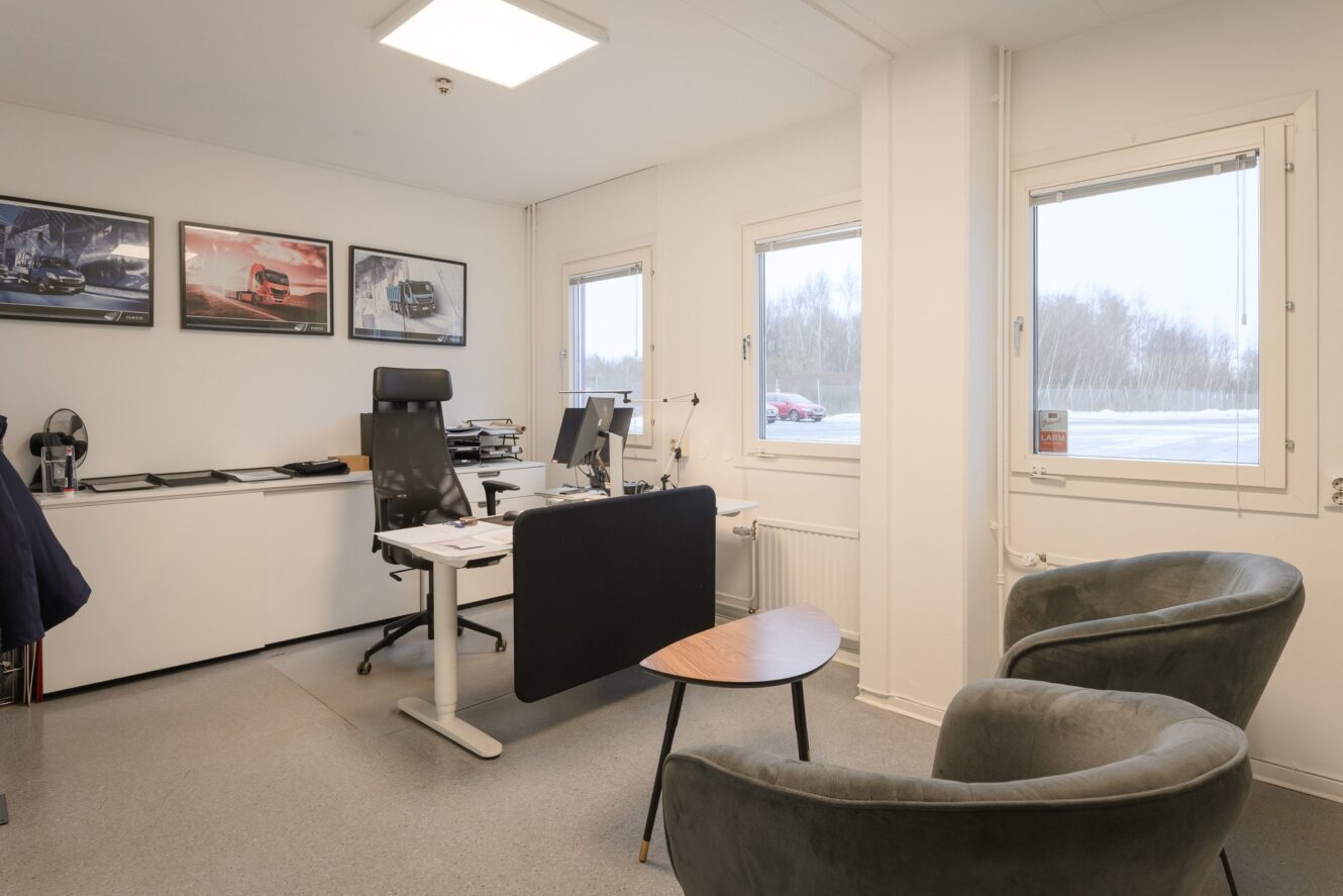 Modernt kontor med skrivbord, stol, dator, två fåtöljer, litet bord och tre tavlor. Stora fönster ger en snöig vy utanför.