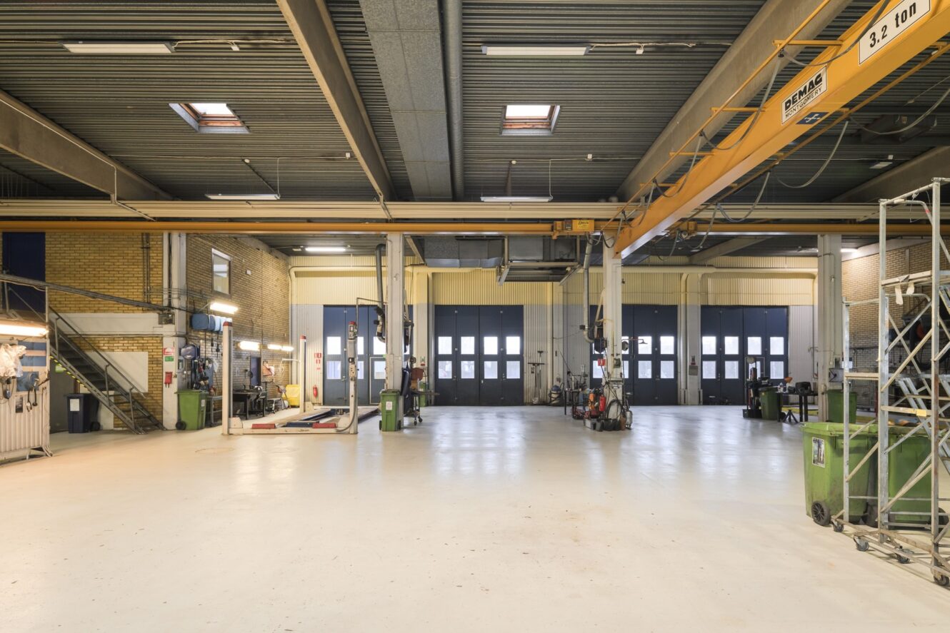 Rymlig industriverkstad med högt i tak, stora garageportar, utrustning, verktyg och en gul kran i ett ljust och rent utrymme.
