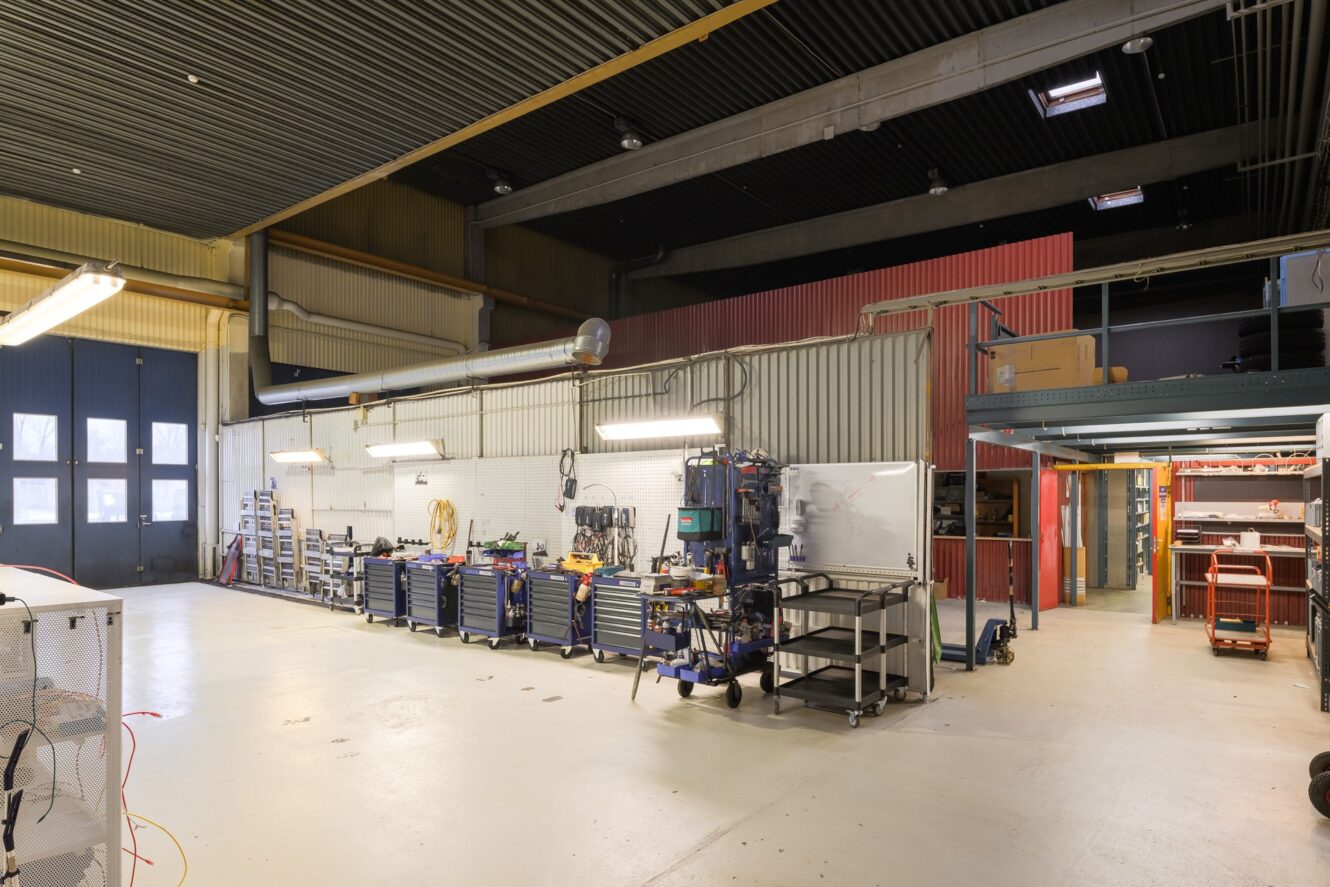 Rymlig industriverkstad med verktyg, arbetsbänkar, förvaringshyllor och utrustning under ett högt tak med stark belysning.