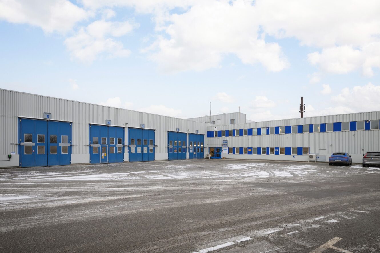 En industribyggnad med blå dörrar och fönster, en stor asfalterad tomt, flera parkerade bilar och lätt snö under en molnig himmel.