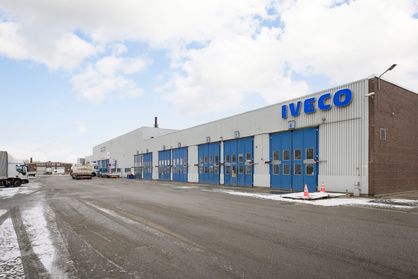 Stor industribyggnad med blå garageportar och IVECO-skylt, på en asfalterad tomt med flera lastbilar och lätt snö som ligger utspridd.