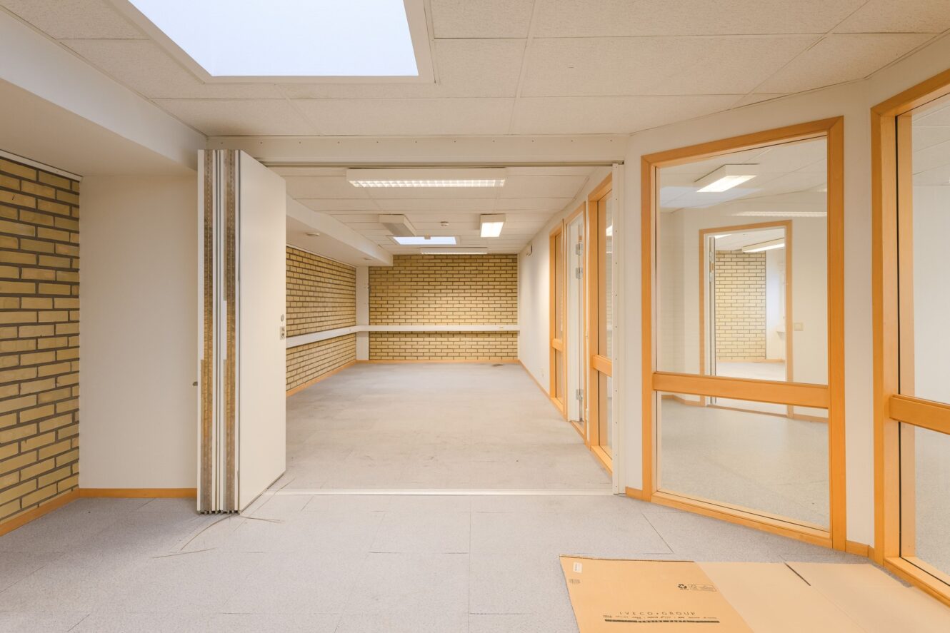 Tomt kontorsutrymme med tegelväggar och vita väggar, glasväggar, taklampor, takfönster och en kartongbit på golvet.