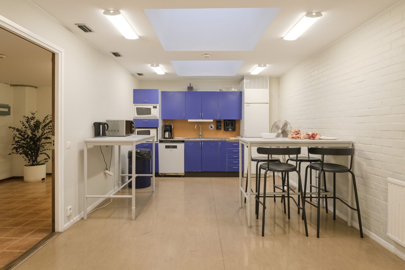 Ett modernt pentry med blå skåp, vita bänkskivor, barbord och pallar, vitvaror, äpplen på ett bord, tegelväggar, takfönster.