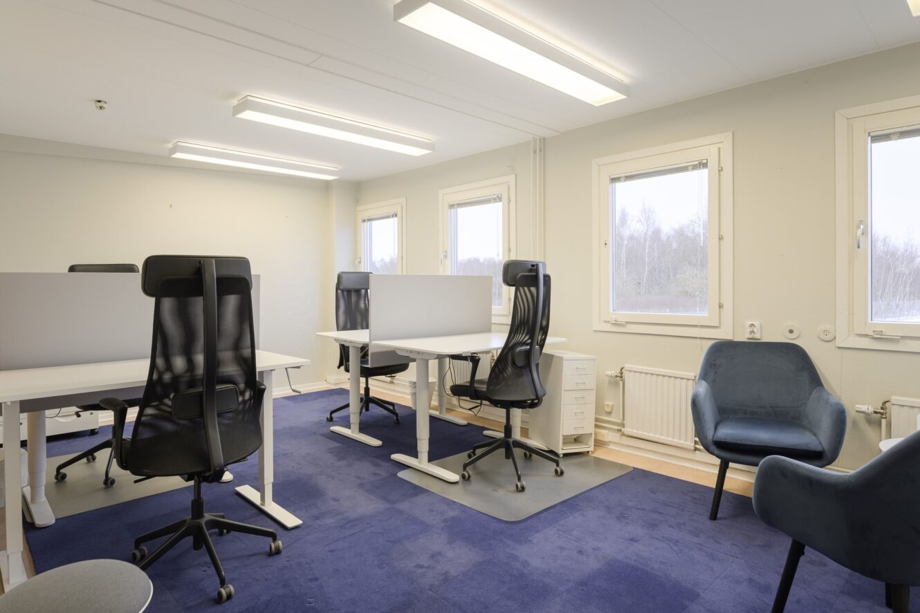 Modernt kontor med skrivbord, svarta ergonomiska stolar, blå matta, två blå fåtöljer, stora fönster och naturligt ljus.