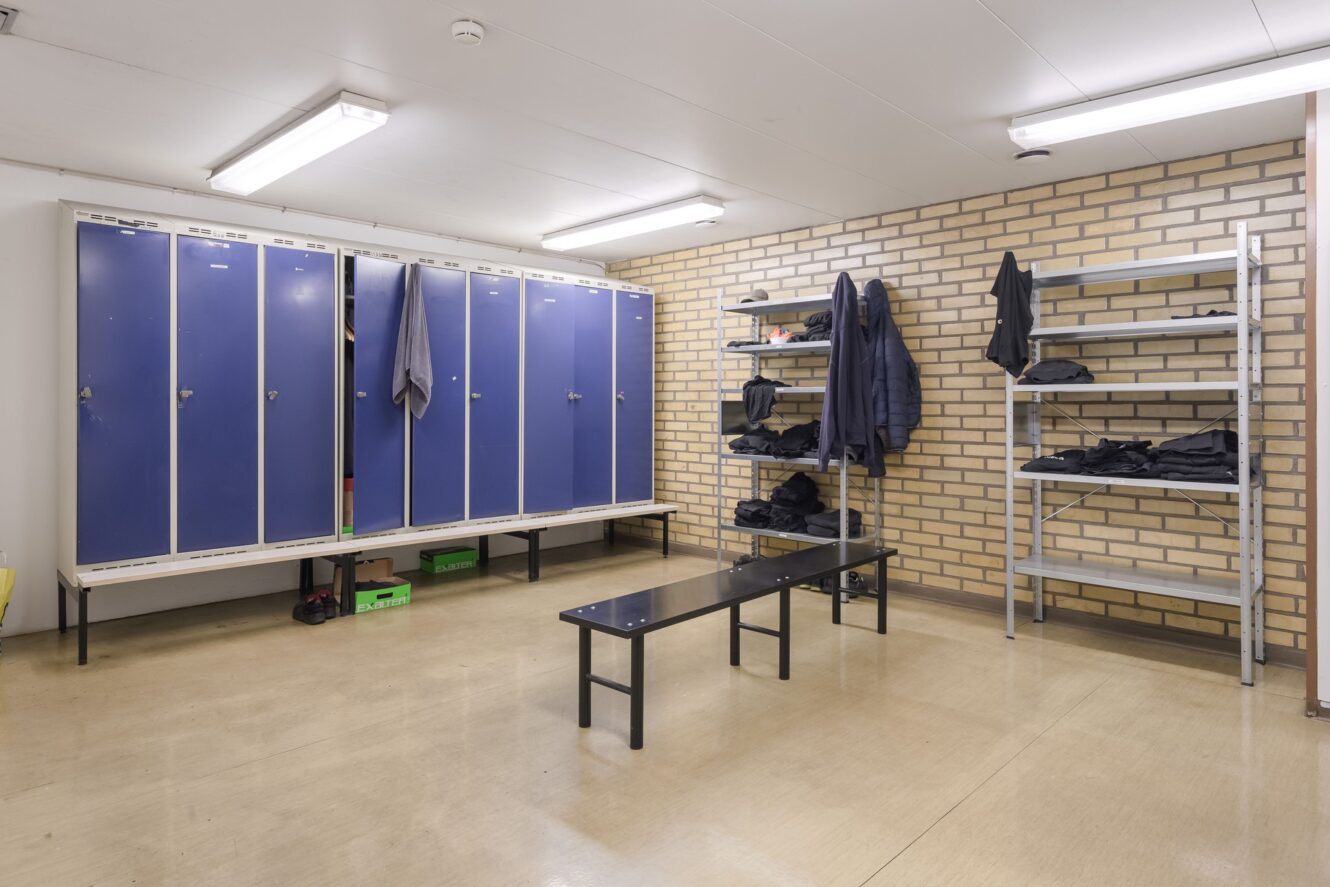Omklädningsrum med blå skåp, metallhyllor med vikta kläder, en bänk i mitten, beige klinkergolv och lysrör i taket.