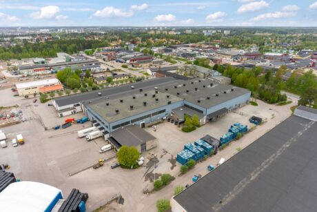 Flygfoto över ett stort industriellt lagerkomplex med lastbilar, fraktcontainrar och närliggande byggnader i ett stadsnära område.