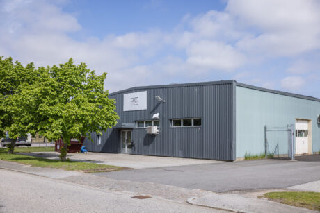 En modern, grå industrilokal med ett litet träd framför, belägen vid en asfalterad gata under en delvis molnig himmel.