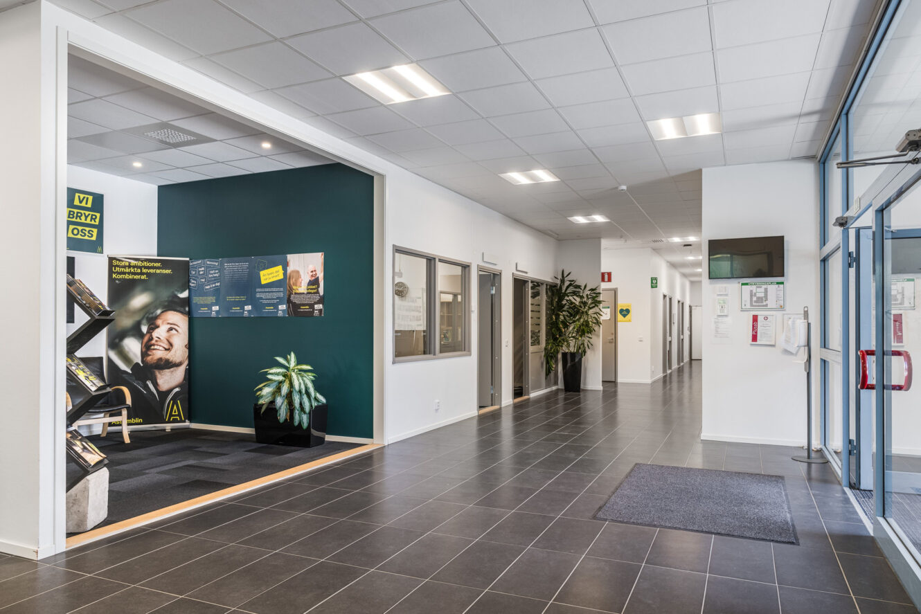 Modern kontorskorridor med klinkergolv, en grön accentvägg med affischer, flera dörrar, krukväxter och en ingång med glasdörr till höger.