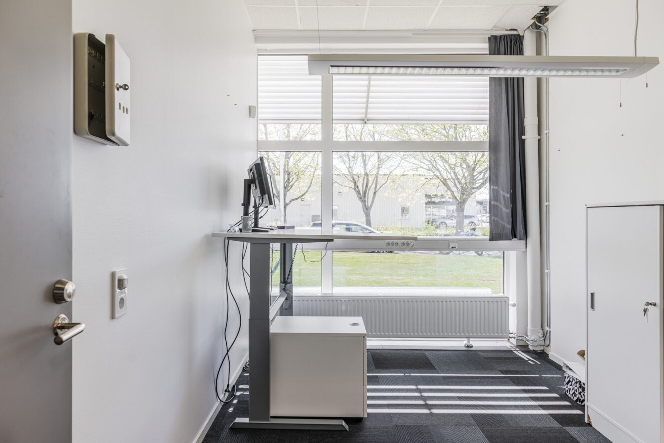 Ett minimalistiskt kontor med ett ståbord, bildskärmar, persienner, ett skåp, gråsvarta mattplattor och träd som syns utanför.