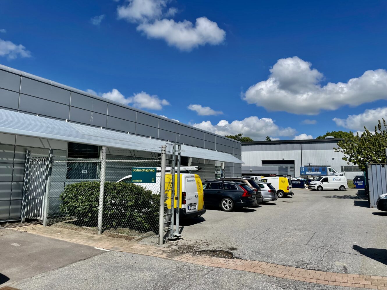 En parkeringsplats med flera bilar och gula skåpbilar bredvid grå industribyggnader under en blå himmel med spridda moln.