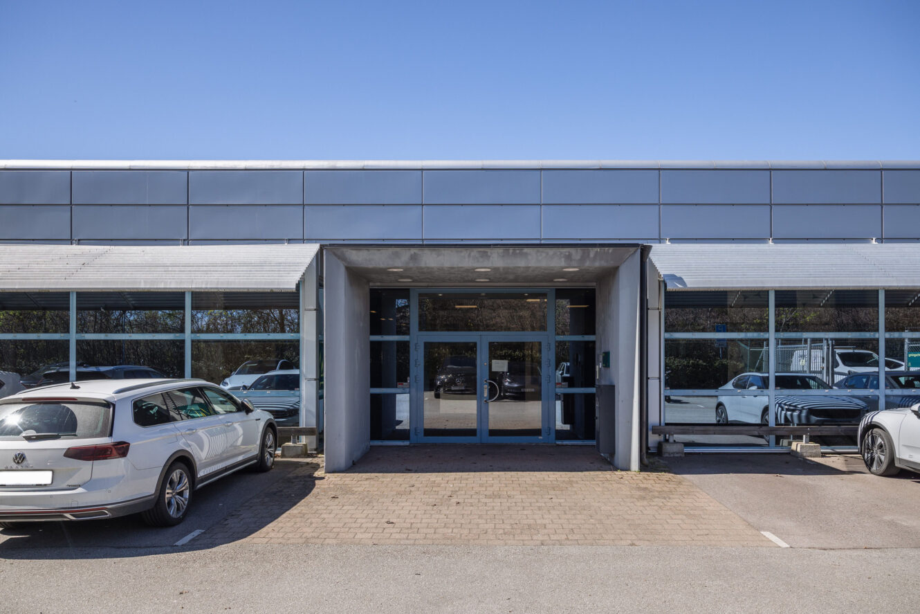 Utsidan av en modern, envånings kontorsbyggnad med stora fönster, dubbla glasdörrar och två bilar parkerade framför byggnaden en klar dag.