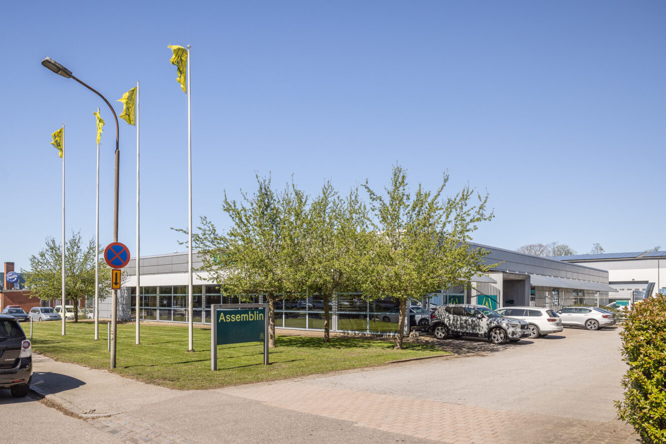 En modern kontorsbyggnad med en grön Assemblin-skylt, gula flaggor, parkerade bilar och träd under en klarblå himmel.