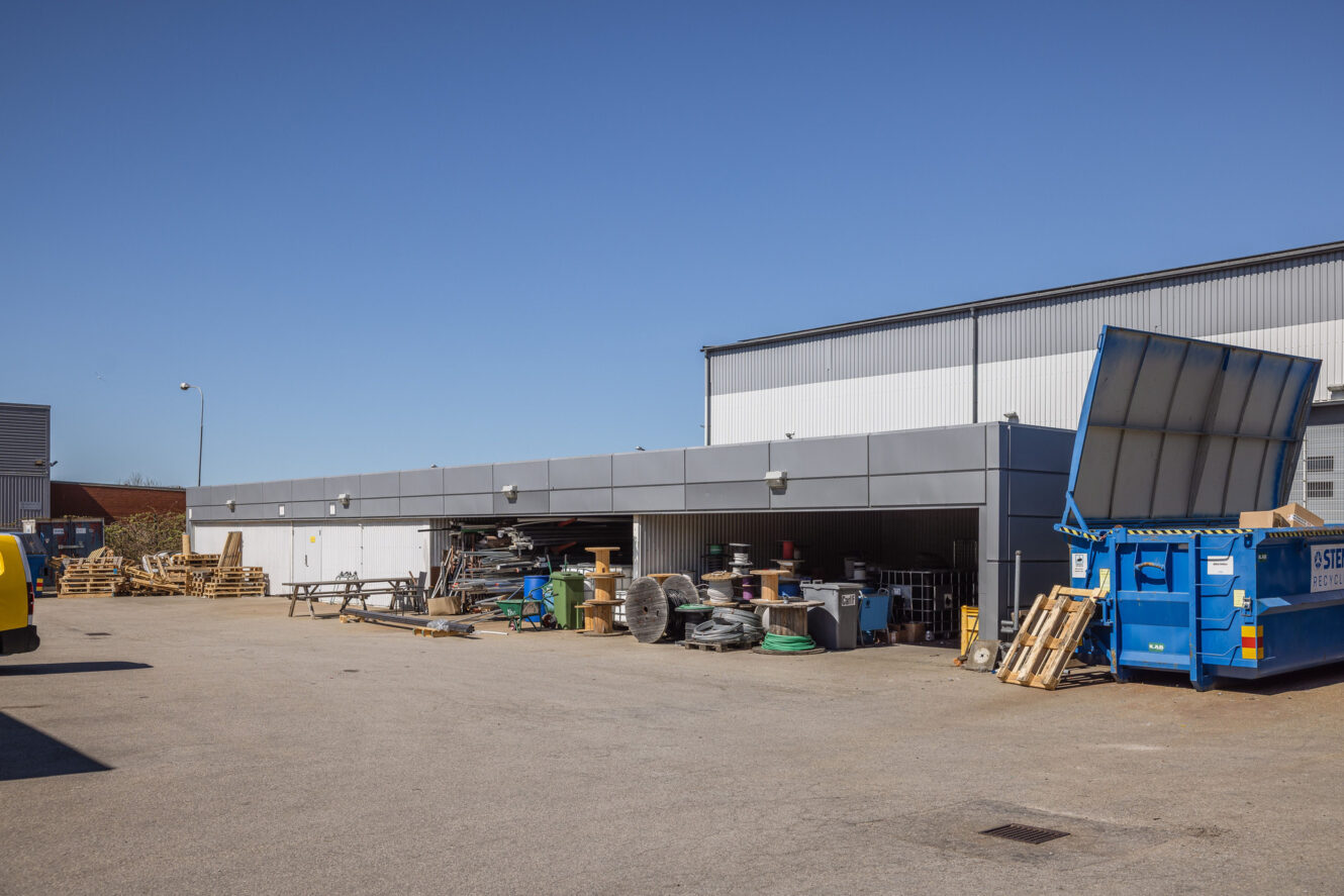 En kommersiell byggnads fasad med staplade pallar, byggmaterial och en blå sopcontainer på en asfalterad tomt under en klar himmel.