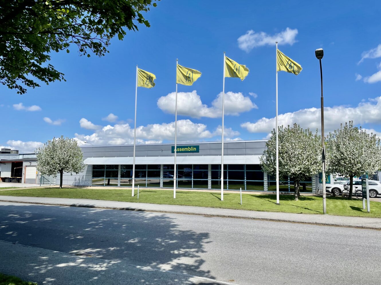 En modern byggnad med en Assemblin-skylt, fyra gula flaggor framför och blommande träd under en solig himmel.
