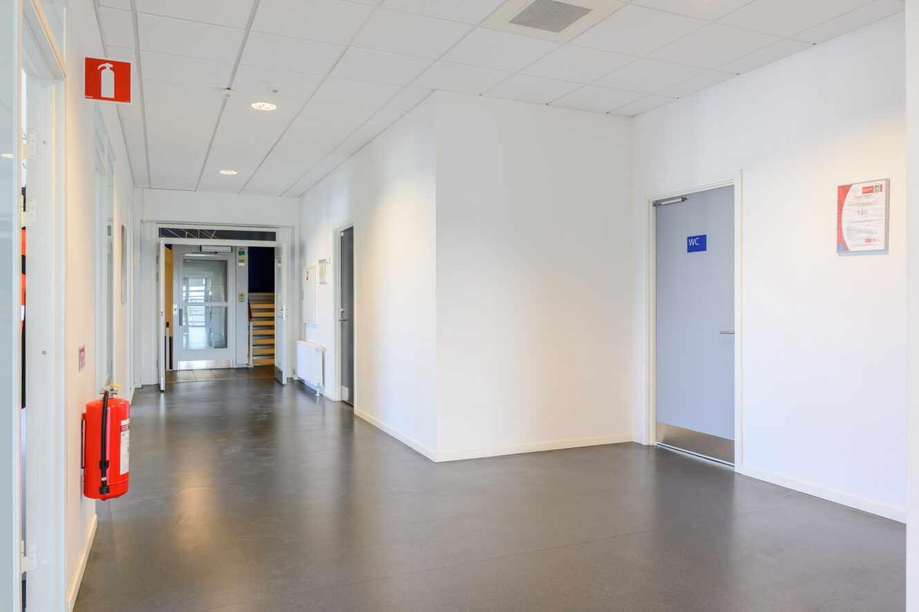 En ren, tom korridor i en byggnad med vita väggar, en synlig brandsläckare, en stängd toalettdörr med skylten ”WC” och taklampor.