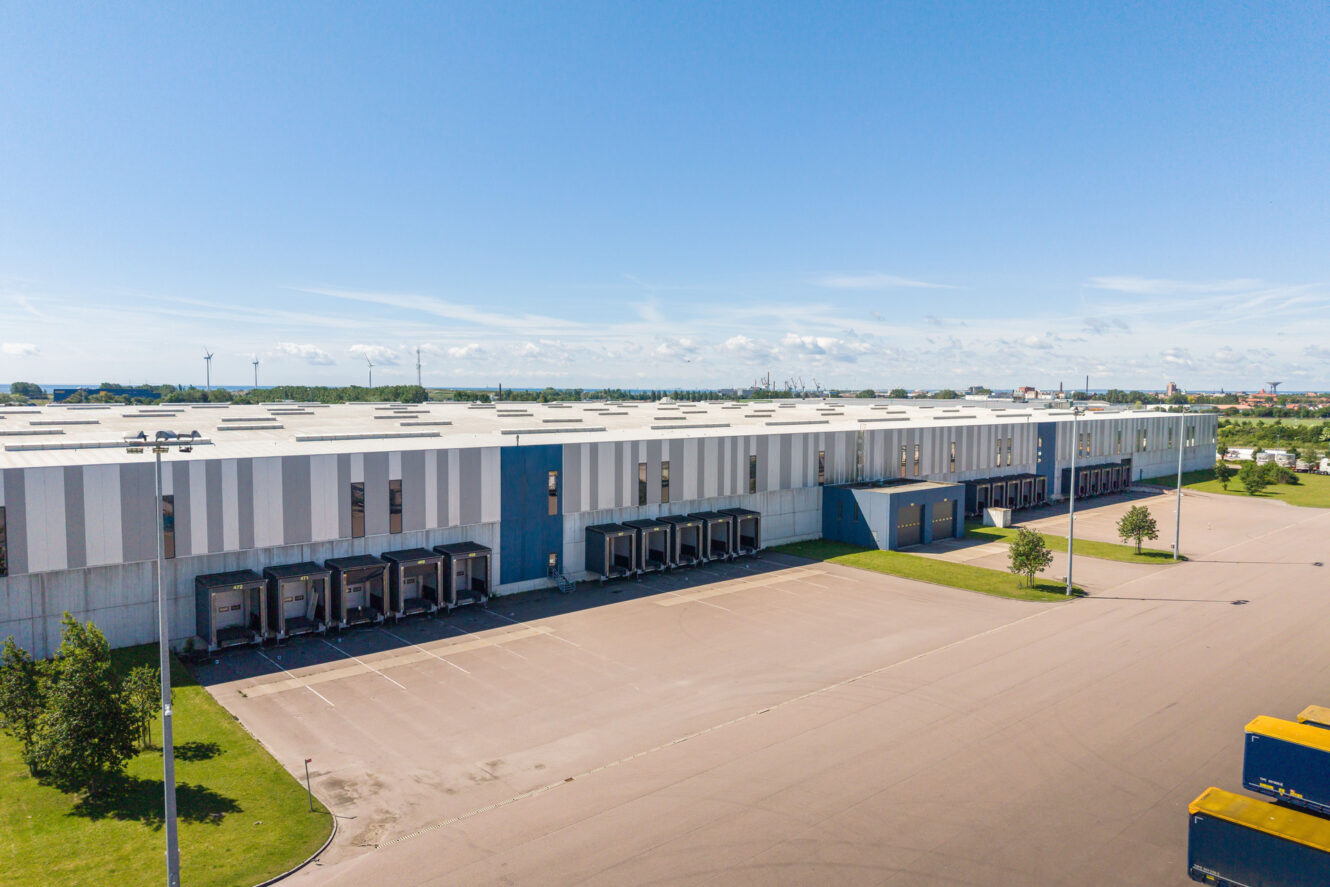 Stort industrilager med flera lastkajer, blå och vit fasad, en tom parkeringsplats och en klarblå himmel i bakgrunden.