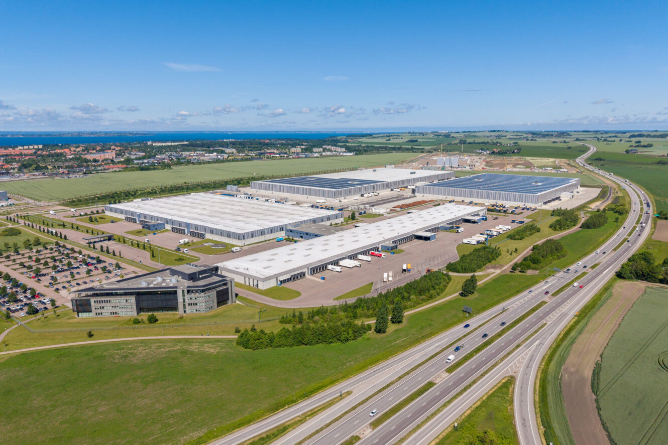 Flygbild över en stor industrianläggning med flera lagerbyggnader med solpaneler på taken, parkeringsplatser, lastbilar och närliggande motorvägar, belägen i ett landsbygdsområde under en klar himmel.