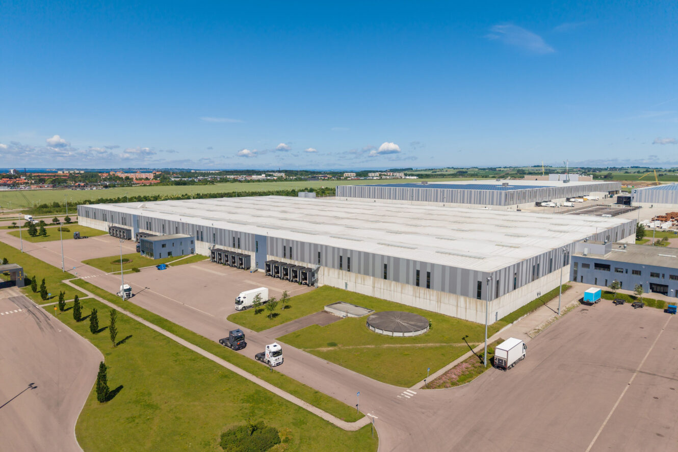 Flygbild över en stor, modern lageranläggning med lastkajer, parkerade lastbilar och omgivande vägar under en klarblå himmel.