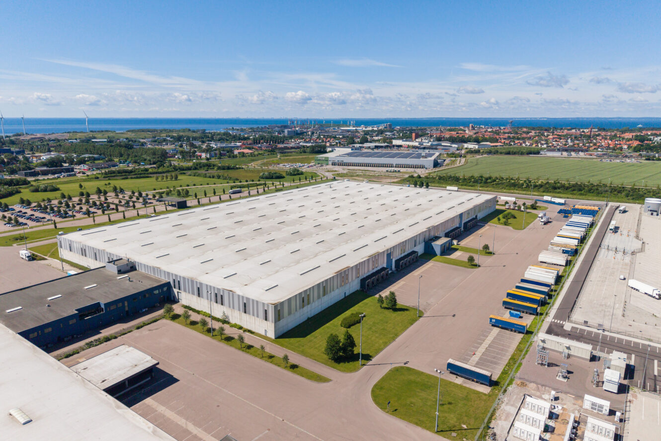 Flygbild över ett stort lager med flera lastkajer och parkerade semitrailers, omgivet av vägar, gräs och närliggande industribyggnader under en klar himmel.