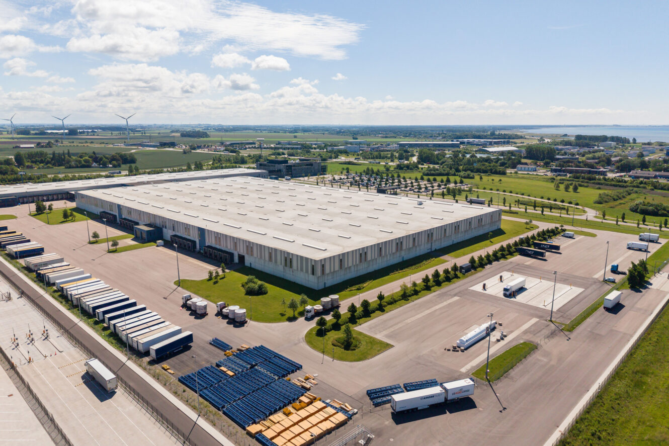 Ett stort lager med flera lastkajer, lastbilar och släpvagnar parkerade utanför, omgivet av gröna fält under en delvis molnig himmel.